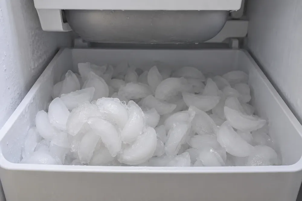 Ice bin of crescent cubes below a freezer ice maker, with several cubes frozen into a clump