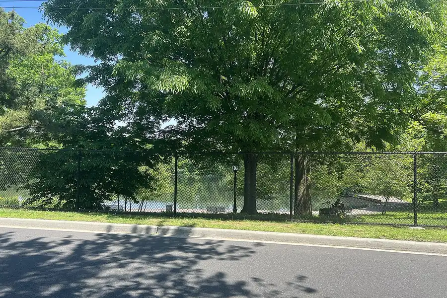 Smith Pond edged by a fence and large shade trees in a park in Roosevelt, New York
